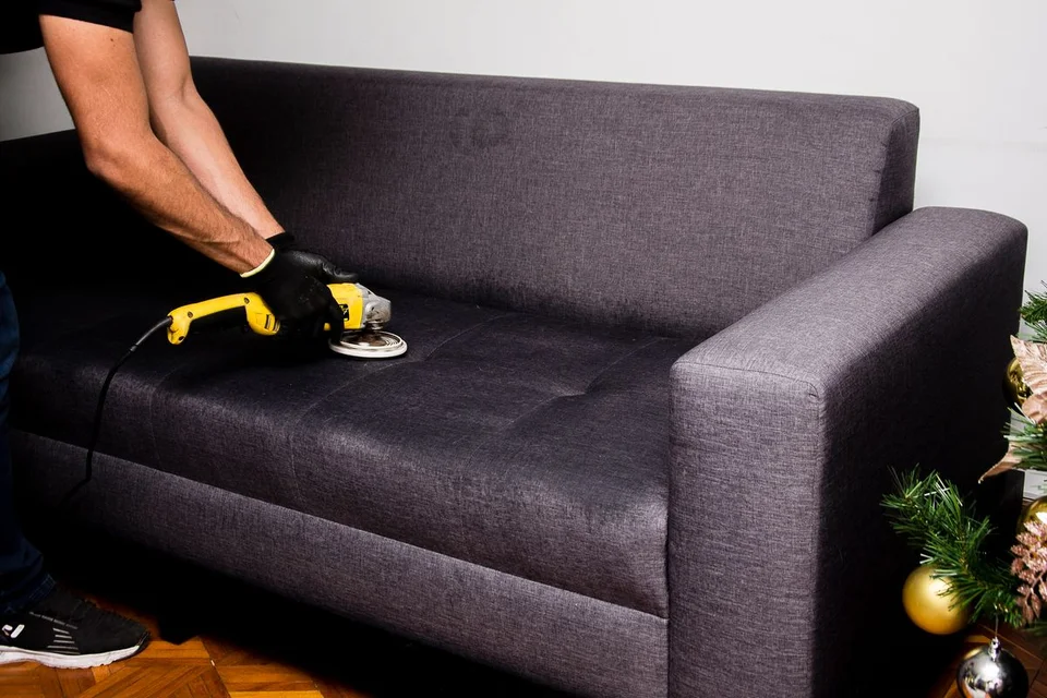 A cleaner using a rotary upholstery brush on the seat of a grey fabric couch